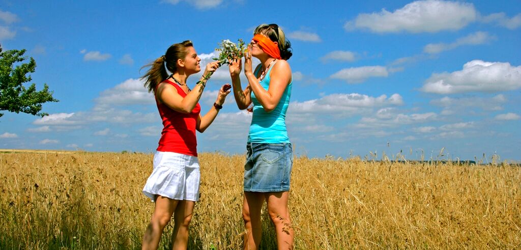 Sommerlich bekleidete Frau steht im Weizenfeld und lässt ihre augenverbundene Freundin an einem Wildblumenstrauß riechen